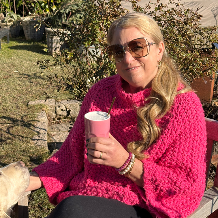 Frau in pinkem grob gestricktem Pullover hält rosa Becher draußen an sonnigem Tag mit Hund neben ihr