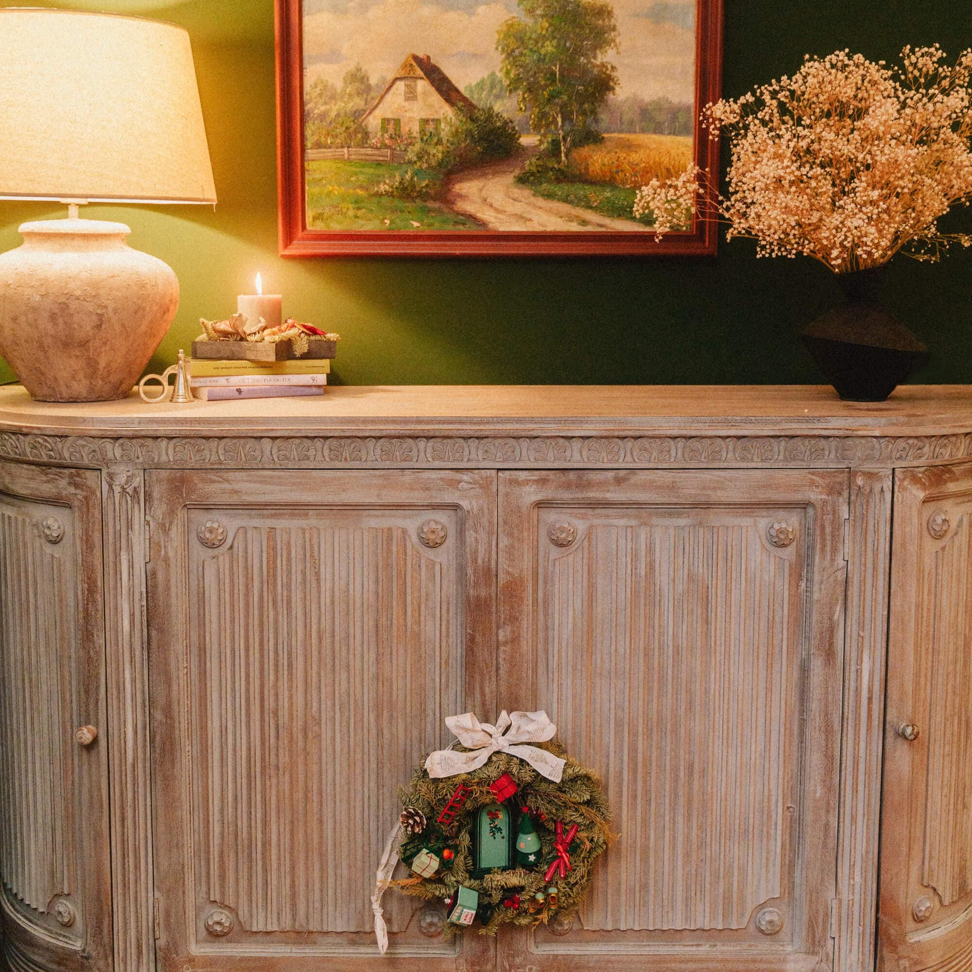Rustikale Holzkommode mit Weihnachtskranz, Lampe und Landschaftsgemälde an grüner Wand