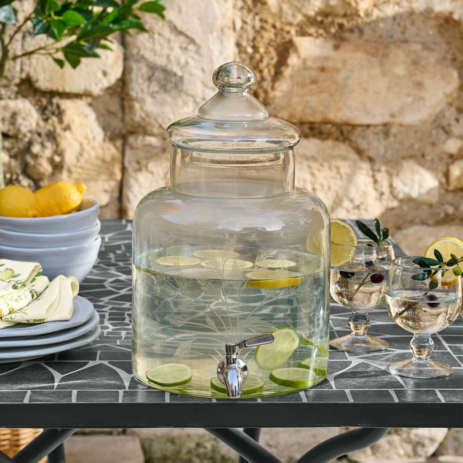 Glasgetränkespender mit Deckel und Zapfhahn, gefüllt mit Zitrone und Limette, auf Gartentisch