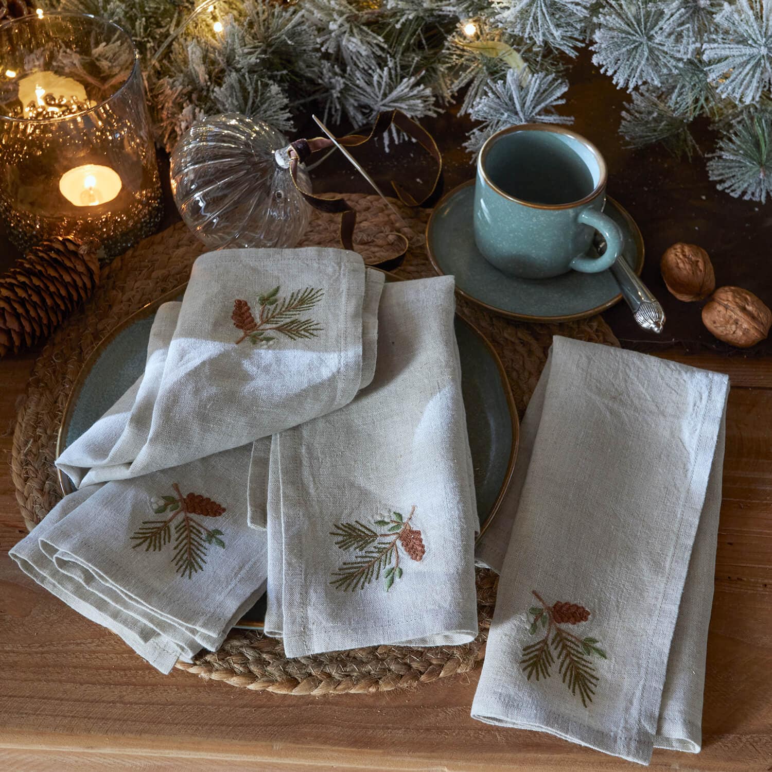 Leinenservietten mit Kiefernzapfen-Stickerei auf rustikalem Tisch mit grünem Becher und Weihnachtsdeko
