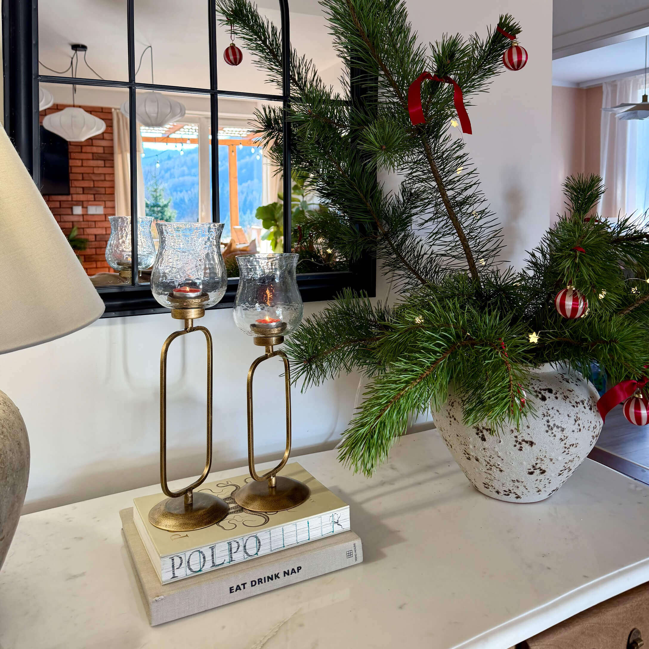 Goldene Kerzenhalter mit klarem Glas auf Büchern neben Vase mit grünen Zweigen und roten Dekorationen