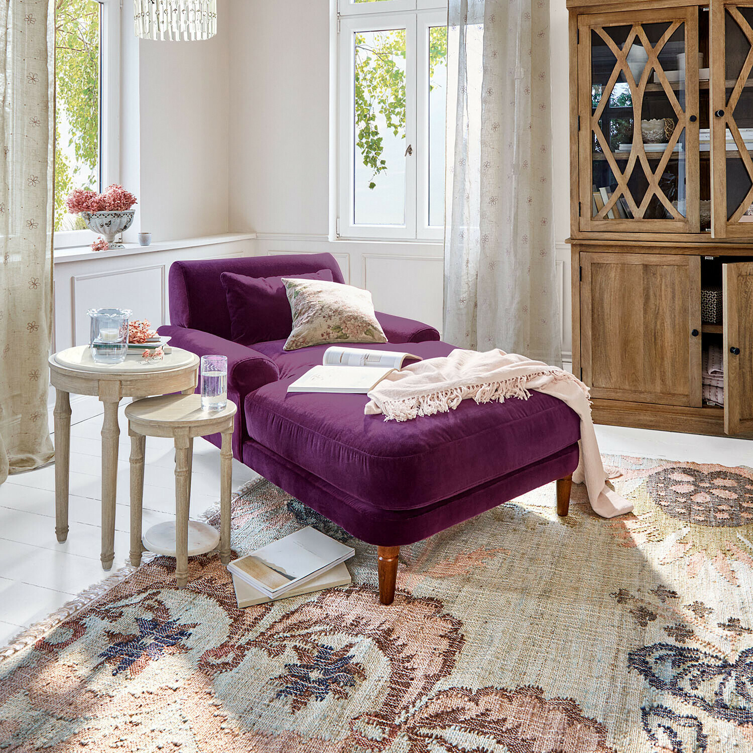 Chaiselounge Jouvrelle Violette Samt-Chaiselongue im hellen Wohnzimmer mit Holzschrank und zwei runden Beistelltischen