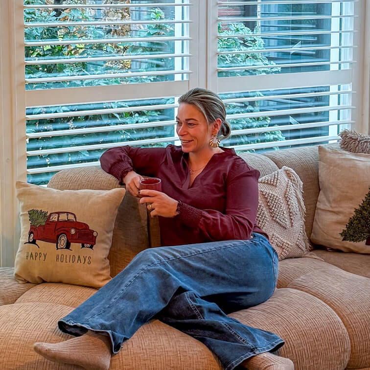 Frau in bordeauxrotem Oberteil und Jeans entspannt auf beigem Sofa mit rotem Weihnachtskissen und Tasse