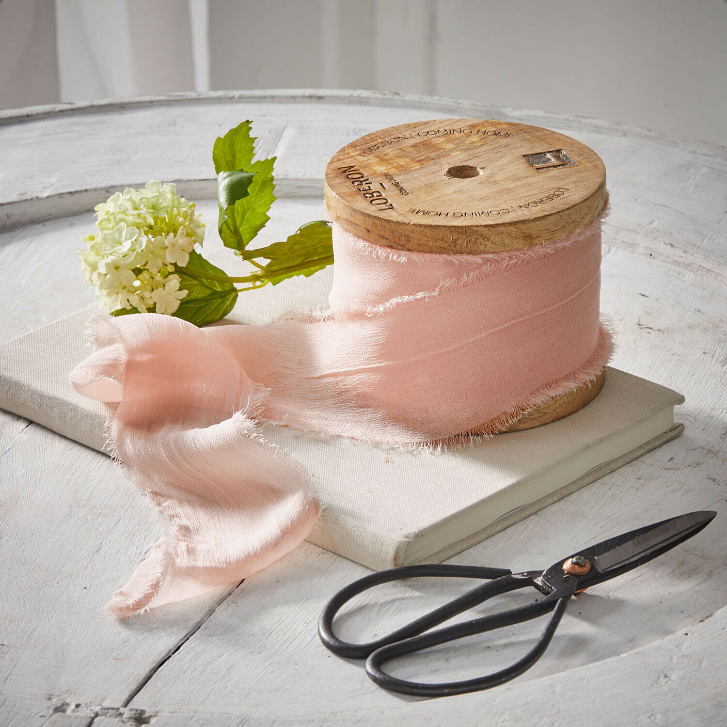 Rosa Stoffband auf Holzspule mit Schere und Blume auf weißem Tisch arrangiert