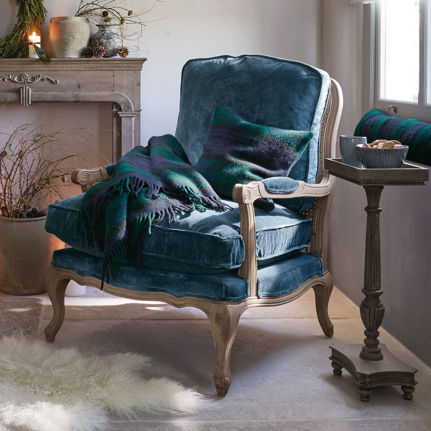 Sessel Corlay Blauer Samtfotel mit hellem Holzrahmen, grün-blauem Plaid und Kissen in gemütlichem Wohnzimmer