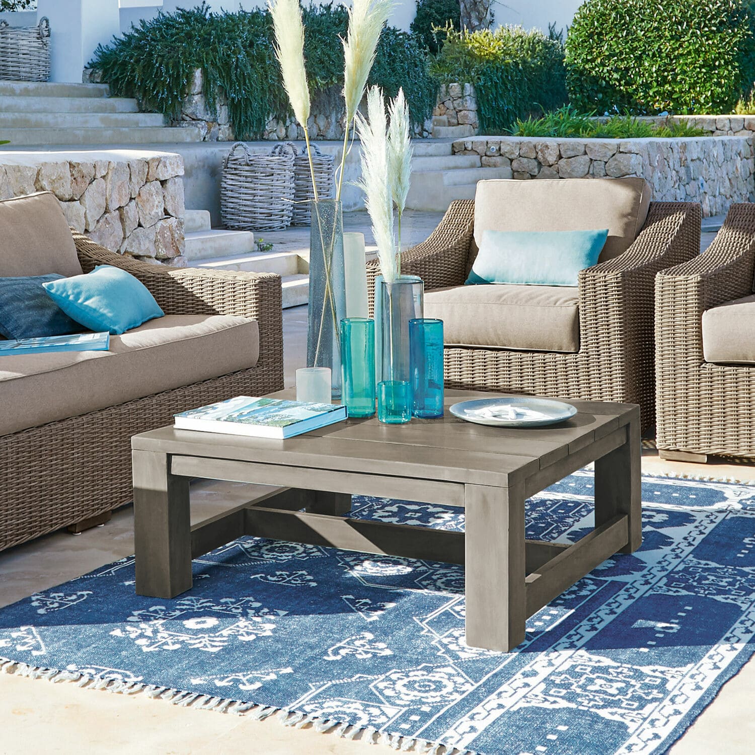 Couchtisch Malden Grauer Holzcouchtisch auf blauem gemustertem Teppich, umgeben von Korbsesseln mit blauen Kissen.