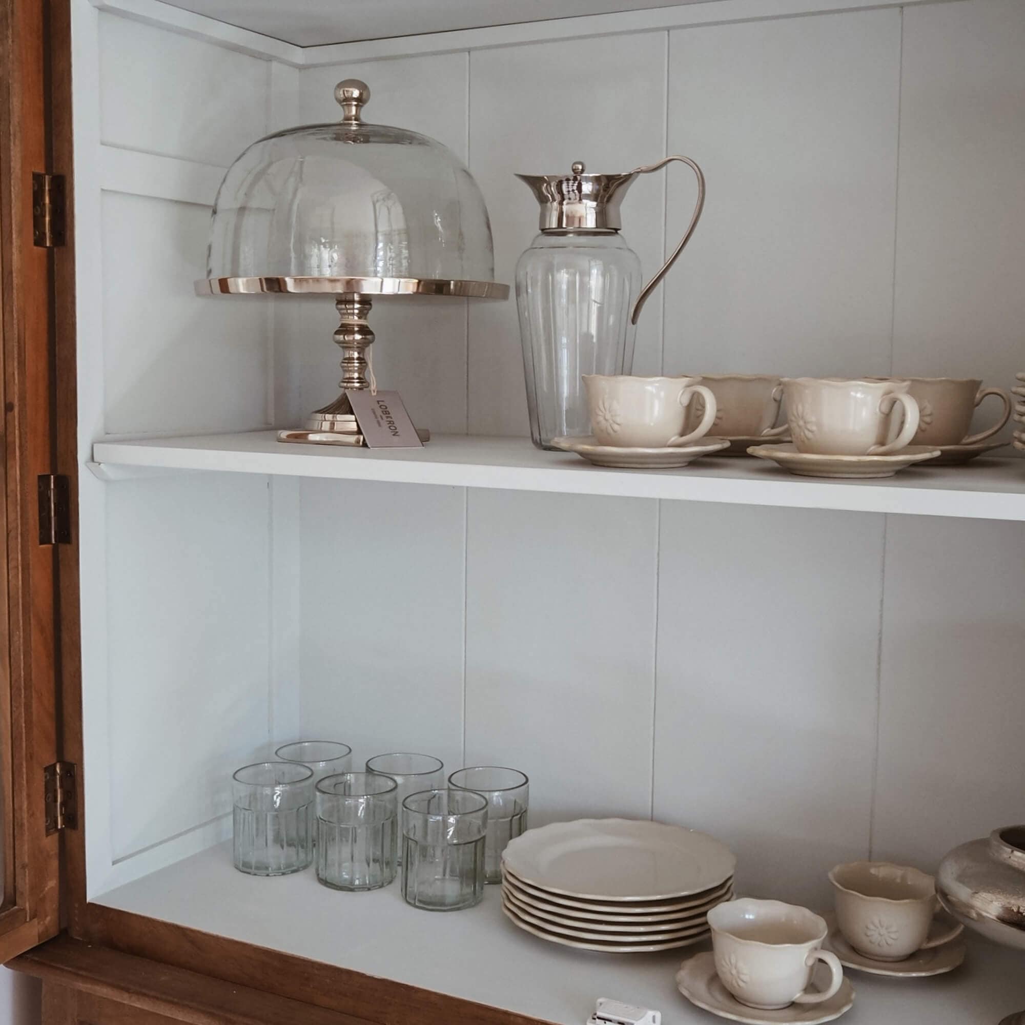 Set aus cremefarbenen Keramiktassen und Tellern mit Glas-Krug und Kuchenhaube in weißem Schrank