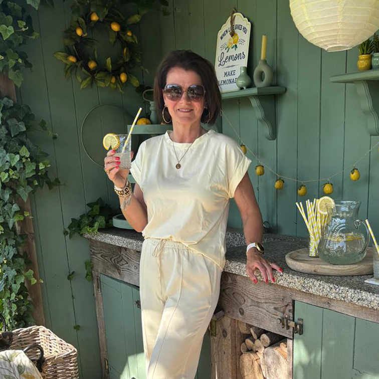 Frau in cremefarbenem Outfit entspannt mit Limonade vor grünem Wandhintergrund mit Zitronendekor