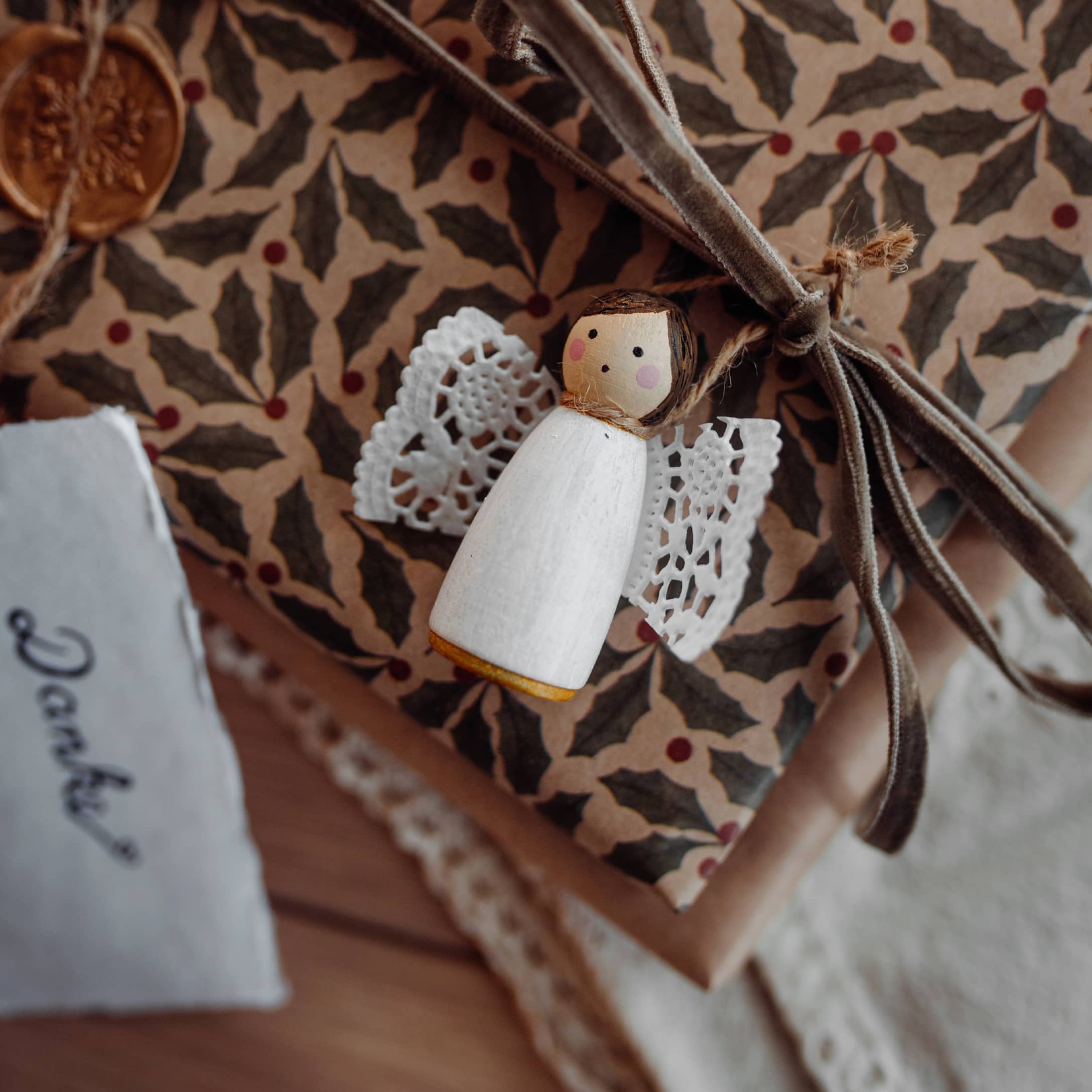Holzengel mit weißen Spitzenflügeln auf Geschenk mit gemustertem Papier und Samtschleife