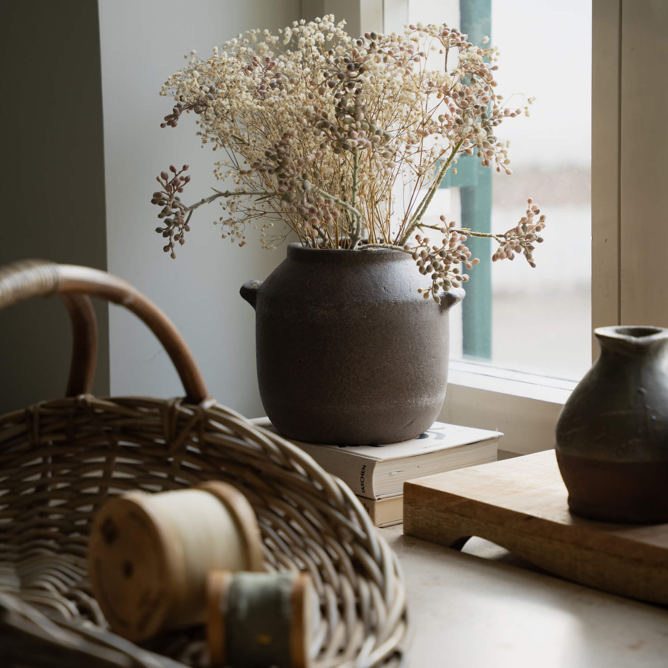 Braune Keramikvase mit getrockneten Blumen auf Fensterbank neben Weidenkorb und Büchern