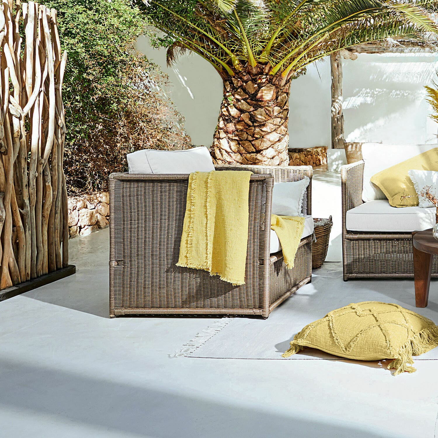 Sessel Lesmont Braunes Rattan-Gartenmöbel-Set mit weißen Kissen und gelben Akzenten auf sonniger Terrasse