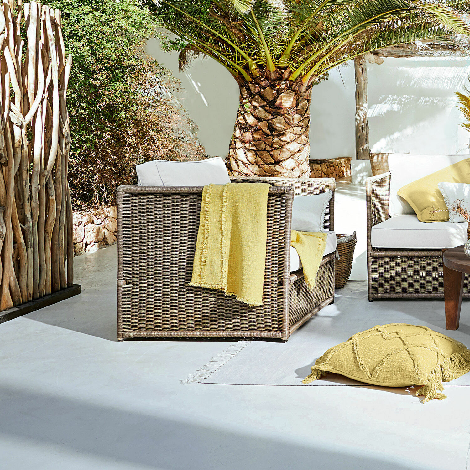 Sessel Lesmont Braunes Rattan-Gartenmöbel-Set mit weißen Kissen und gelben Akzenten auf sonniger Terrasse