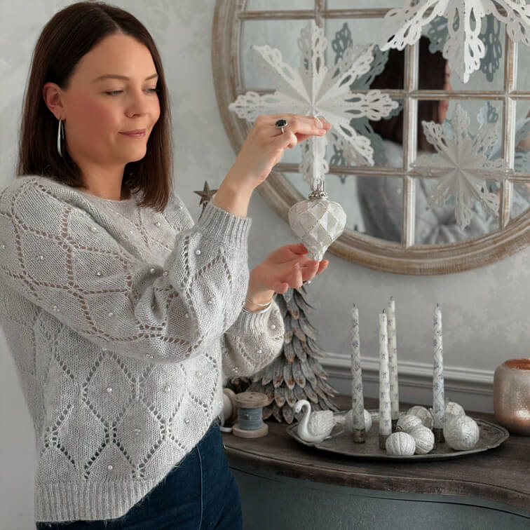 Frau in grauem Perlenstrickpullover hält weiße Weihnachtsdekoration in gemütlichem Raum mit Winterdeko