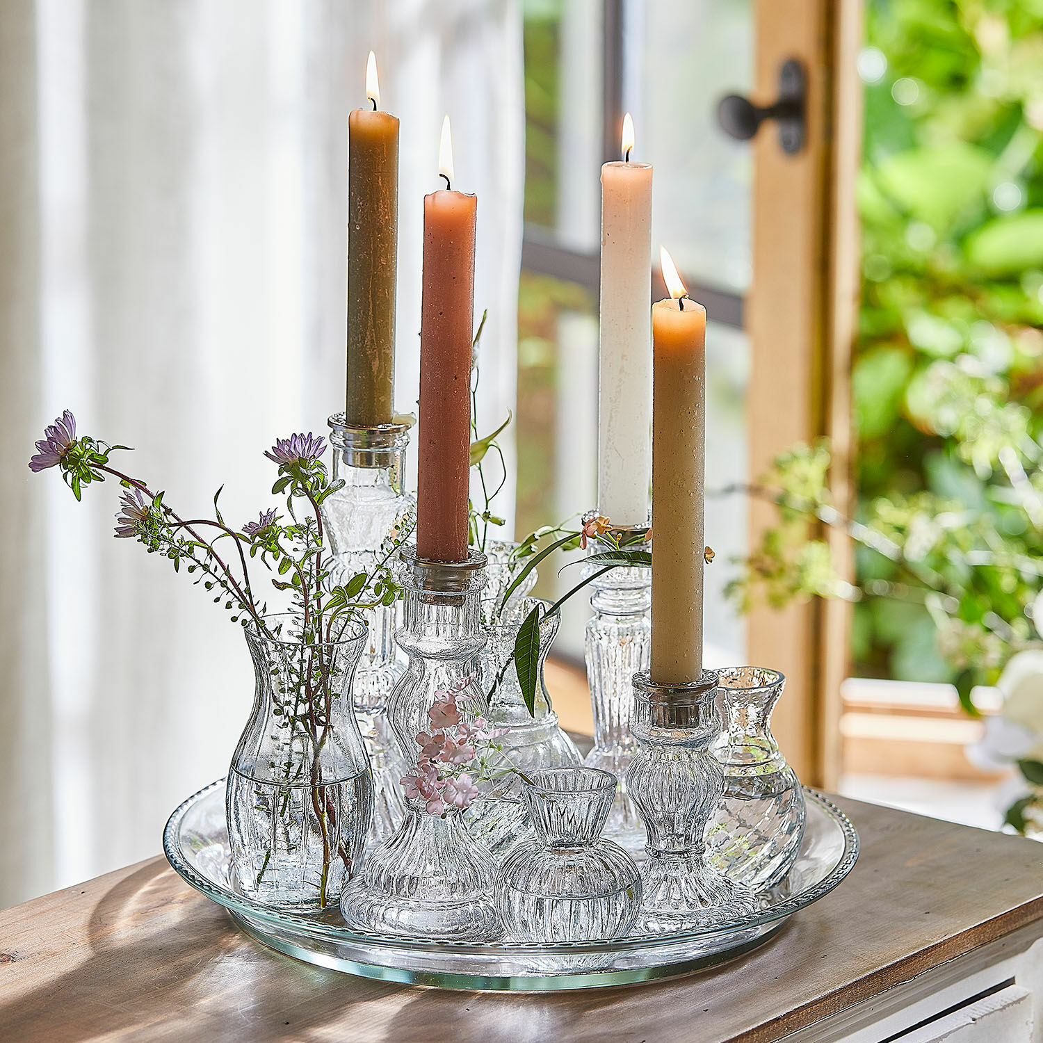 Kerze 12er Set Bauceau Gläserne Kerzenhalter und Väschen mit farbigen Kerzen und Blumen auf Glastablett auf Holztisch