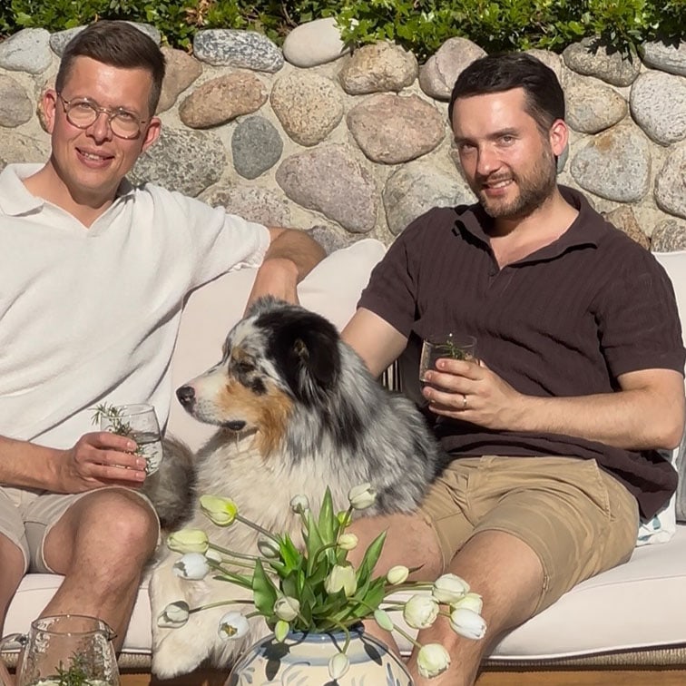 Zwei Männer sitzen draußen auf einer Couch mit einem Australian Shepherd Hund und weißen Tulpen im Vordergrund.