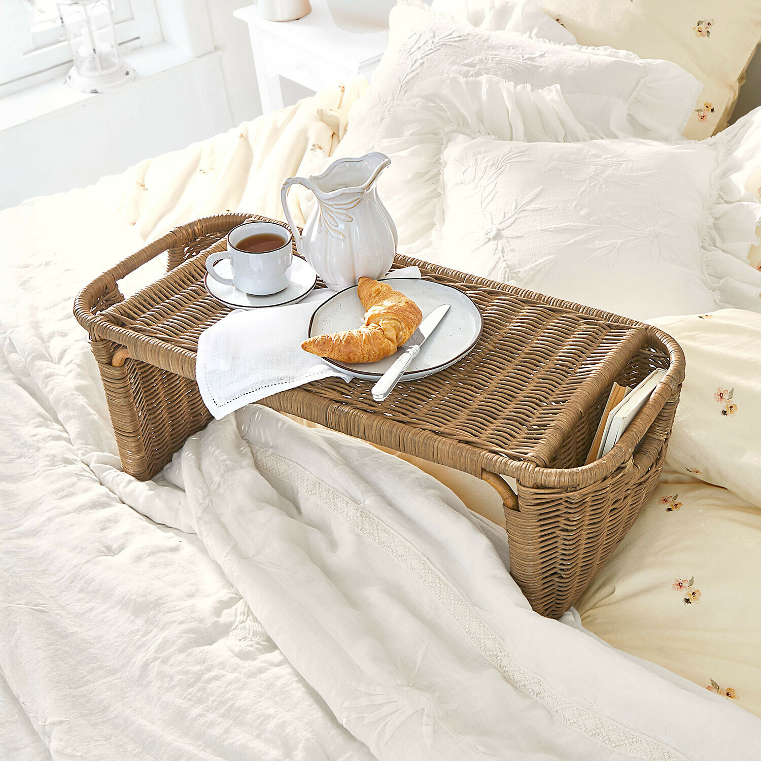 Brauner Rattantablett mit weißem Geschirr und Croissant auf hellem Bett in gemütlichem Schlafzimmer
