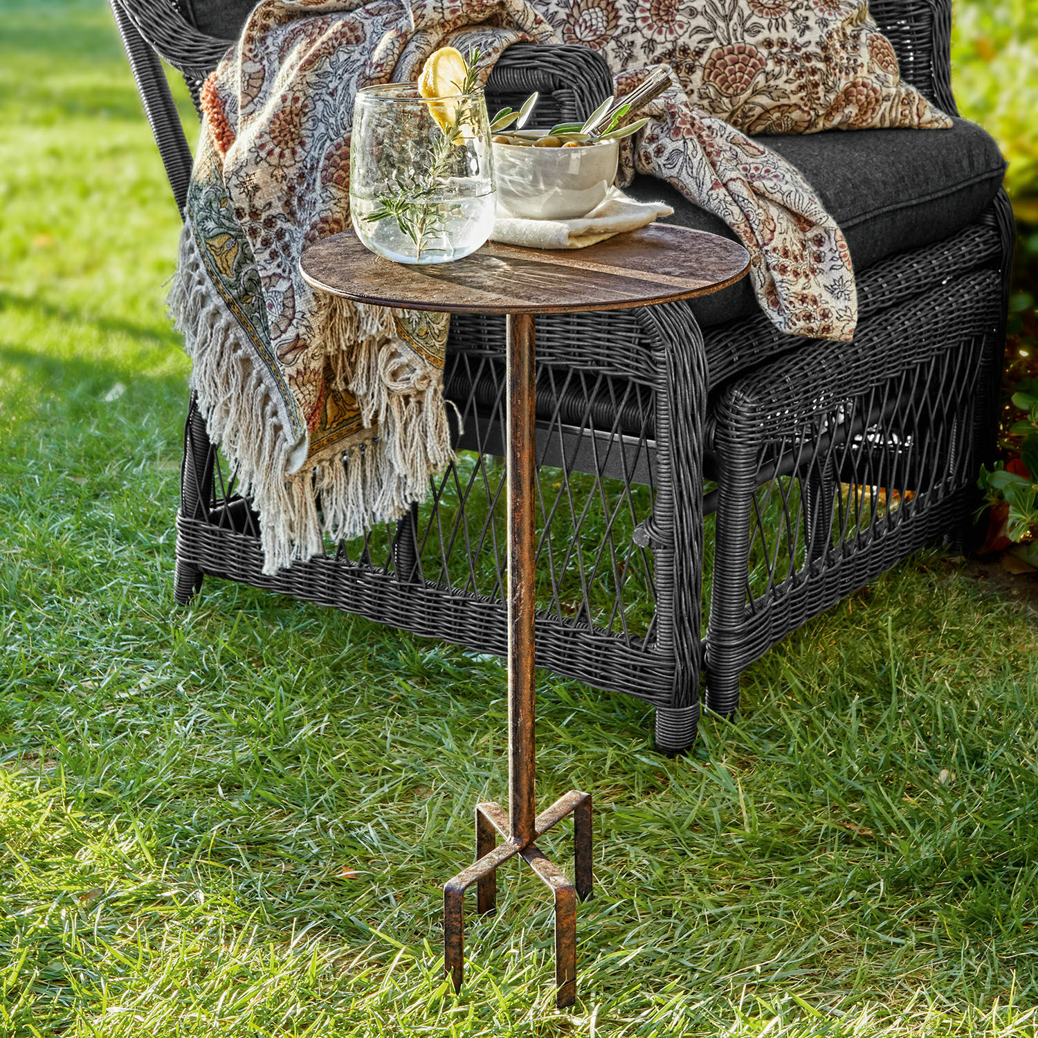 Tischchen Fermont Metall-Beistelltisch mit Vintage-Patina auf Gras neben einem Korbsessel mit Kissen und Decke