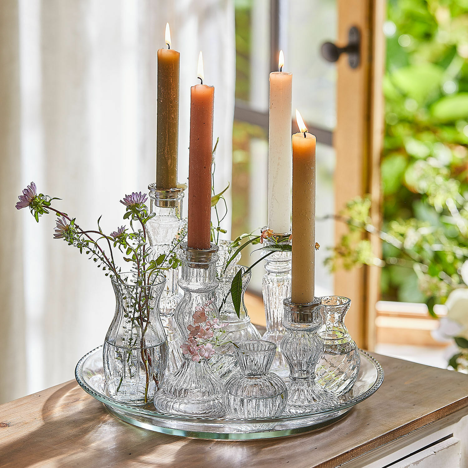 Tablett mit Kerzenständer und Vase Hilevor Gläserne Kerzenhalter und Väschen mit farbigen Kerzen und Blumen auf einem Glastablett auf Holztisch