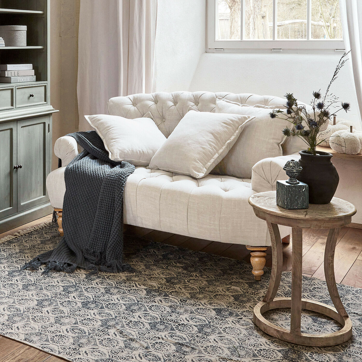 Sofa Springfield Village Beiges, gesteppte Leinen-Sofa mit Kissen und grauer Strickdecke in gemütlichem Wohnzimmer mit Holztisch