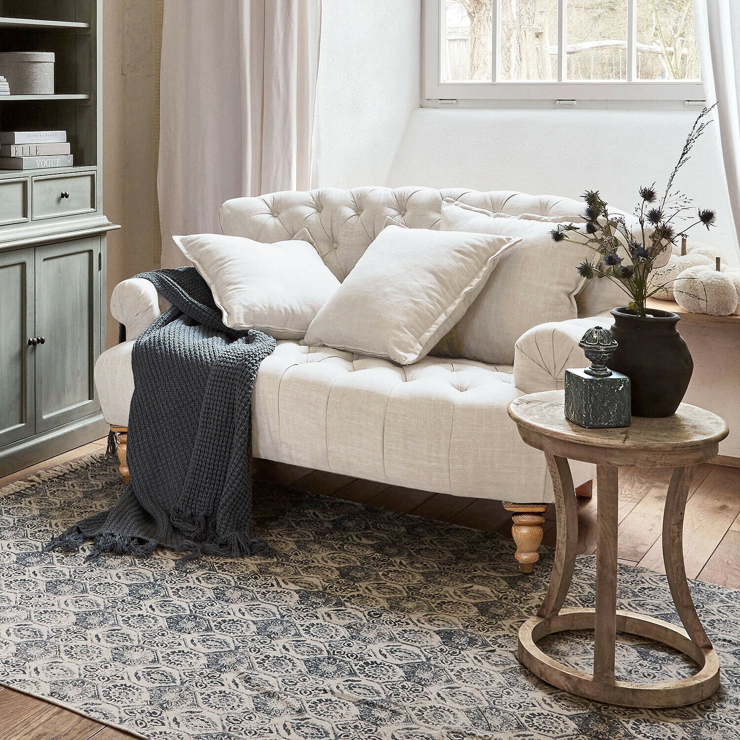 Sofa Springfield Village Beiges, gesteppte Leinen-Sofa mit Kissen und grauer Strickdecke in gemütlichem Wohnzimmer mit Holztisch
