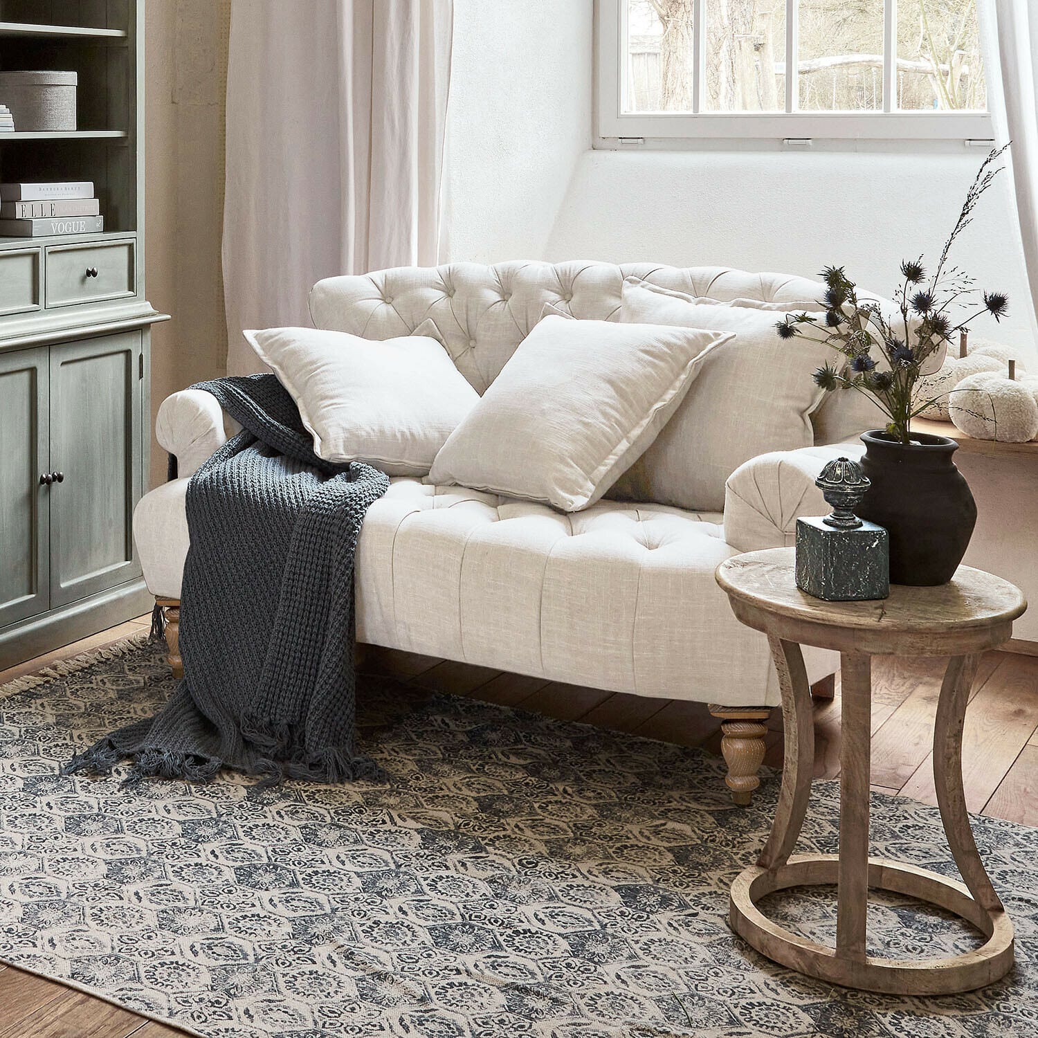 Sofa Springfield Village Beiges, gesteppte Leinen-Sofa mit Kissen und grauer Strickdecke in gemütlichem Wohnzimmer mit Holztisch