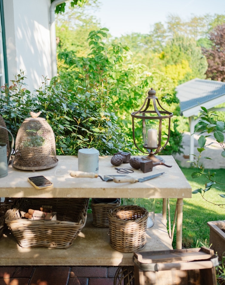 Gartentisch aus Stein mit Weidenkörben, Gartenwerkzeugen und Kerzenlaterne im Grünen