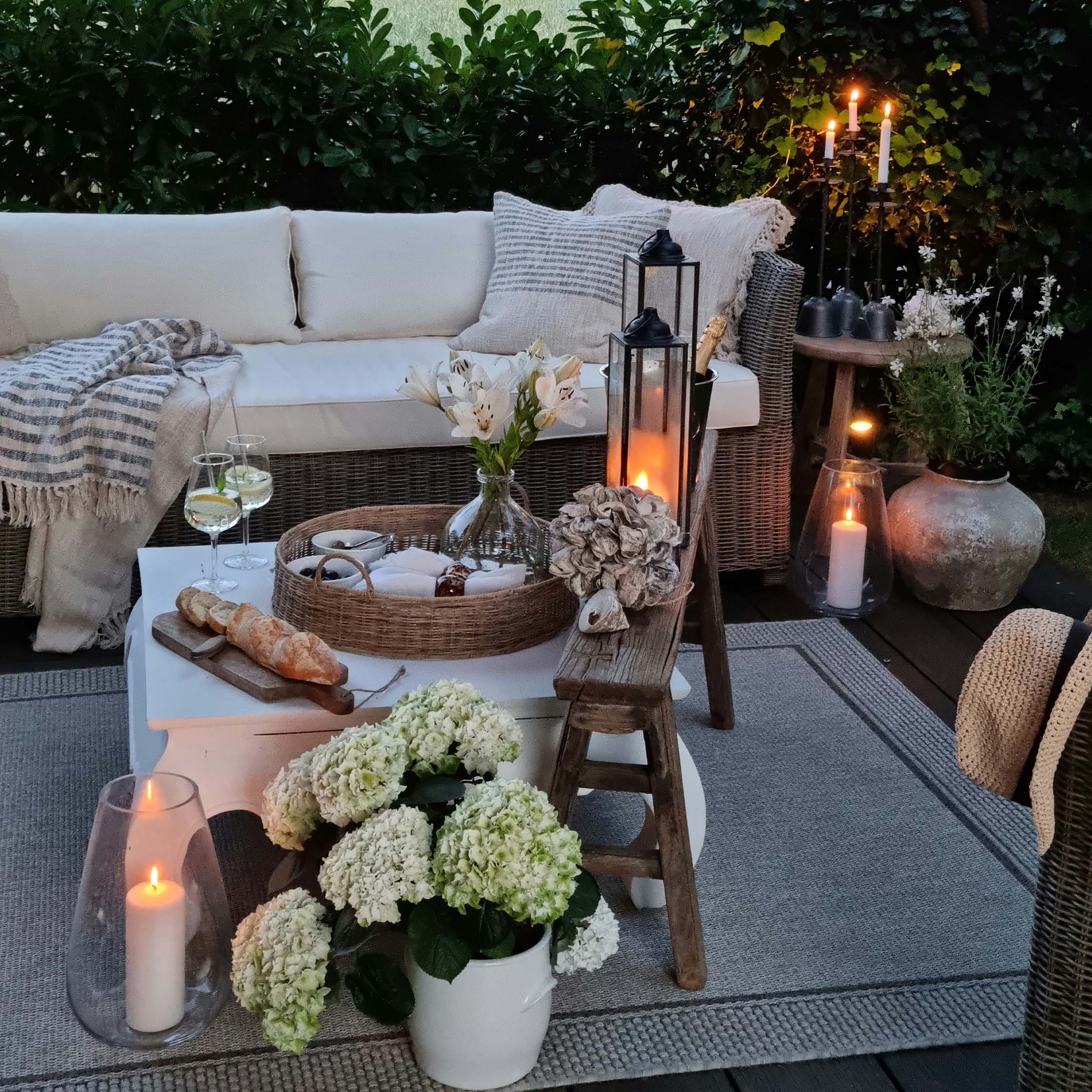 Gemütliche Gartenecke mit Rattansofa, weißen Kissen und Kerzen auf Holzterrasse