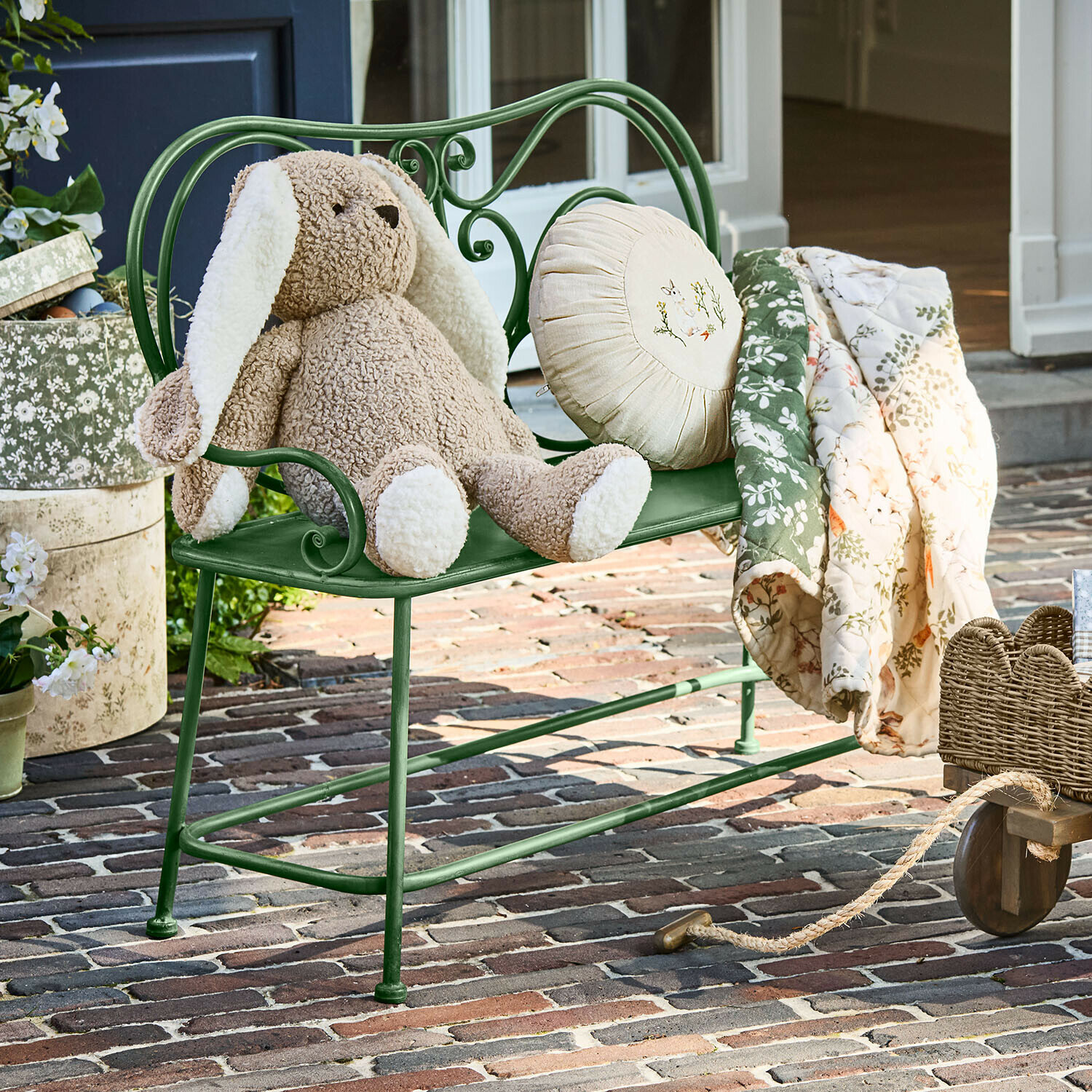 Grüne Metallbank mit beigem Plüschhasen und geblümter Decke auf gepflastertem Hof