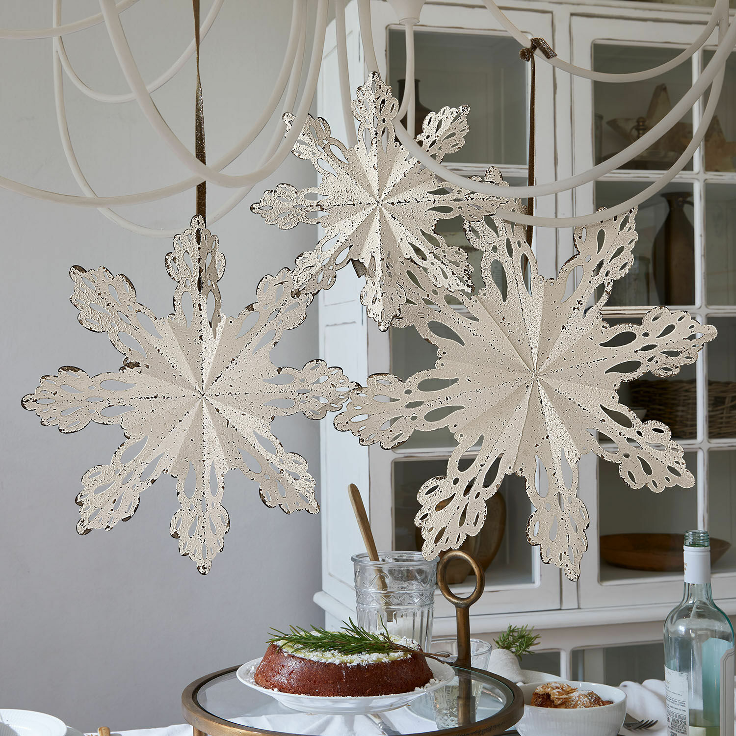 Deko-Ornament 3er Set Lirvaya Dekorative cremefarbene Metall-Schneeflocken hängen in hellem Raum über einem Tisch mit Kuchen