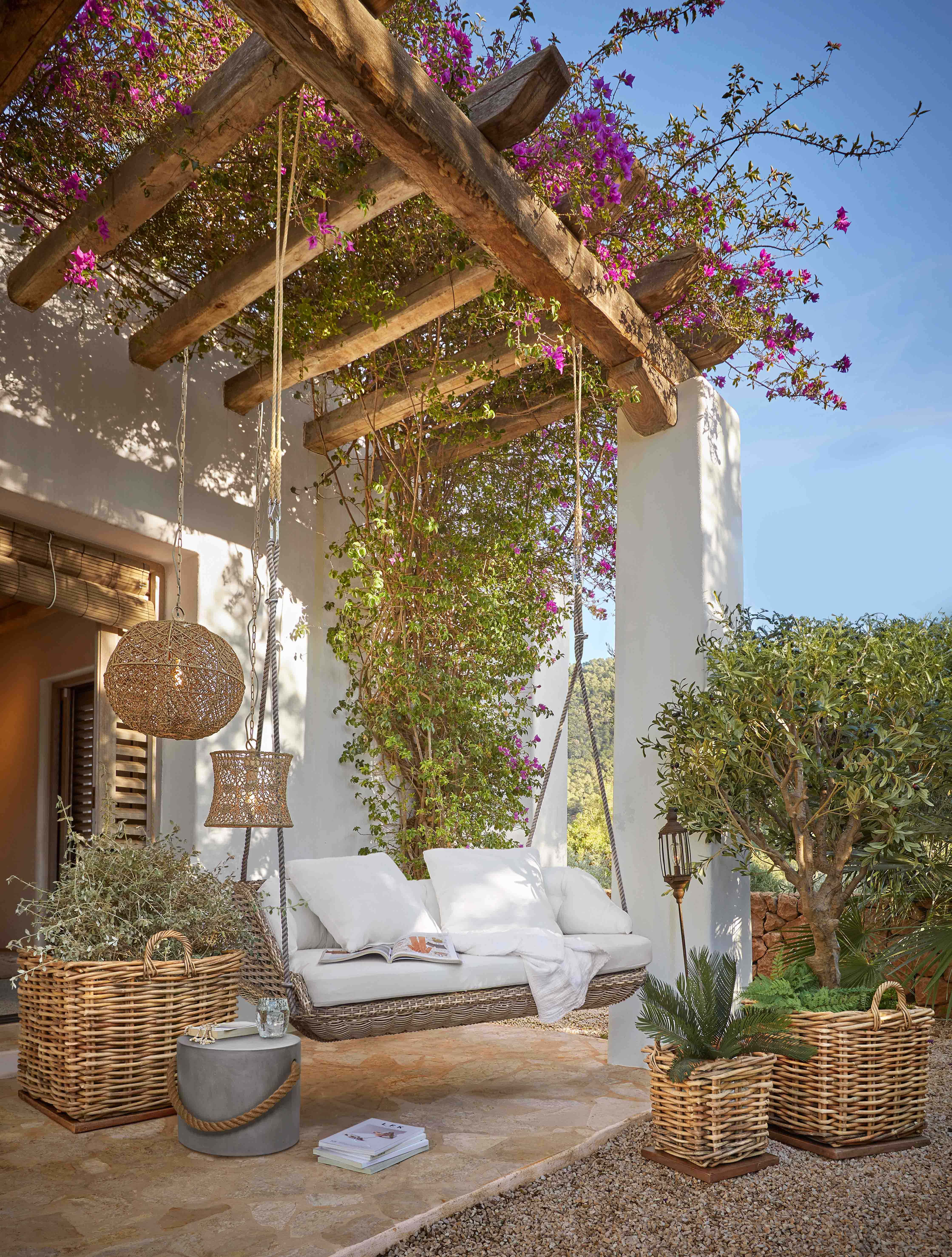 Wikling-Schaukel mit weißen Kissen unter einer Holzpergola mit blühenden pinken Blumen