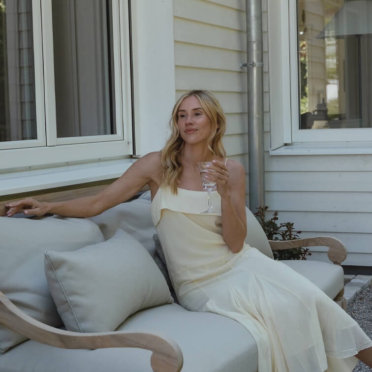 Frau im cremefarbenen Kleid sitzt entspannt auf hellgrauem Sofa draußen und hält ein Weinglas