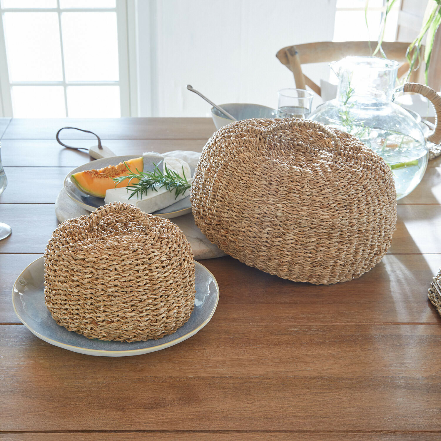 Zwei runde Brotabdeckungen aus geflochtenem Naturmaterial in Beige auf hellem Holztisch