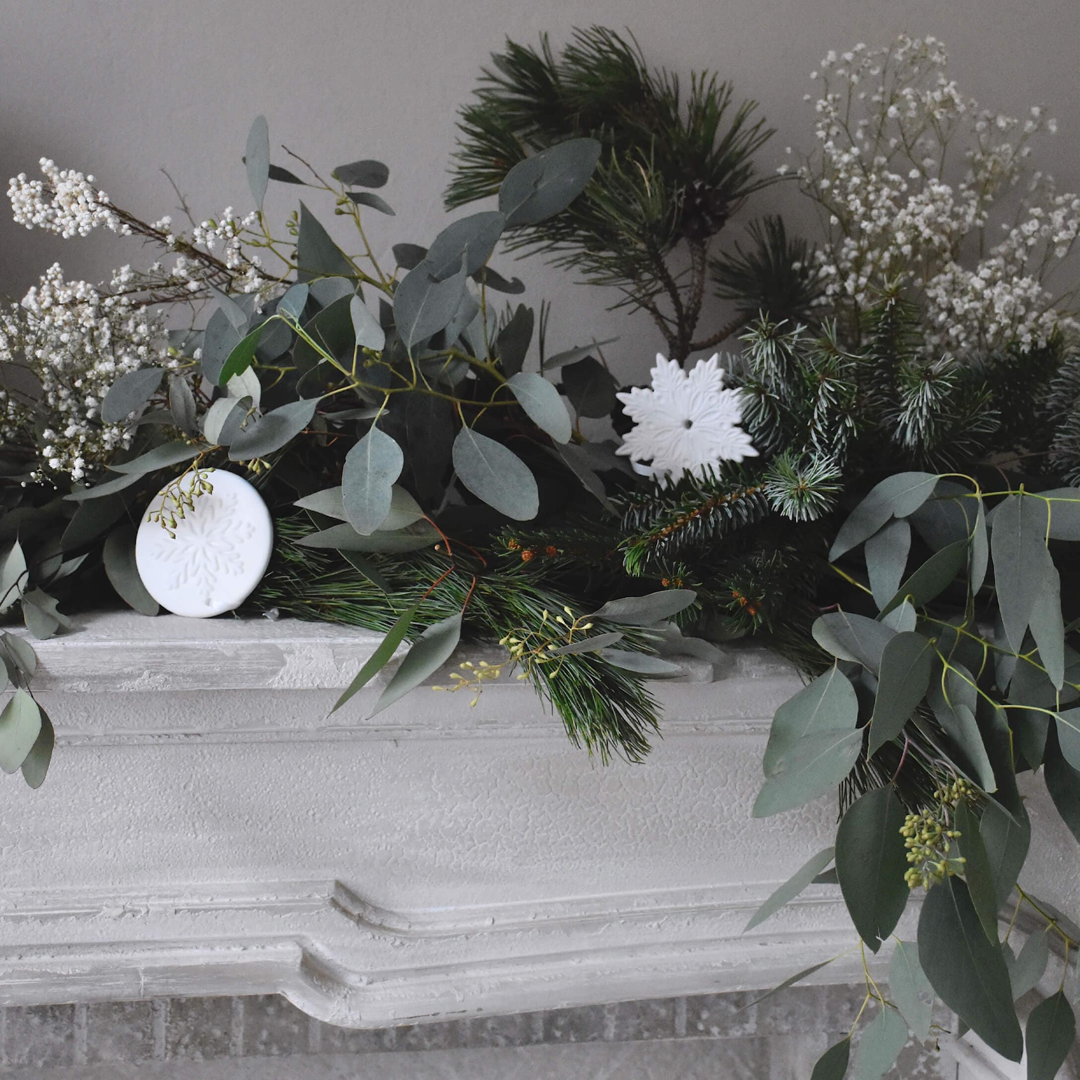 Dekorativer Kranz aus grünen Zweigen und weißen Schneeflockenornamenten auf weißem Steinkamin