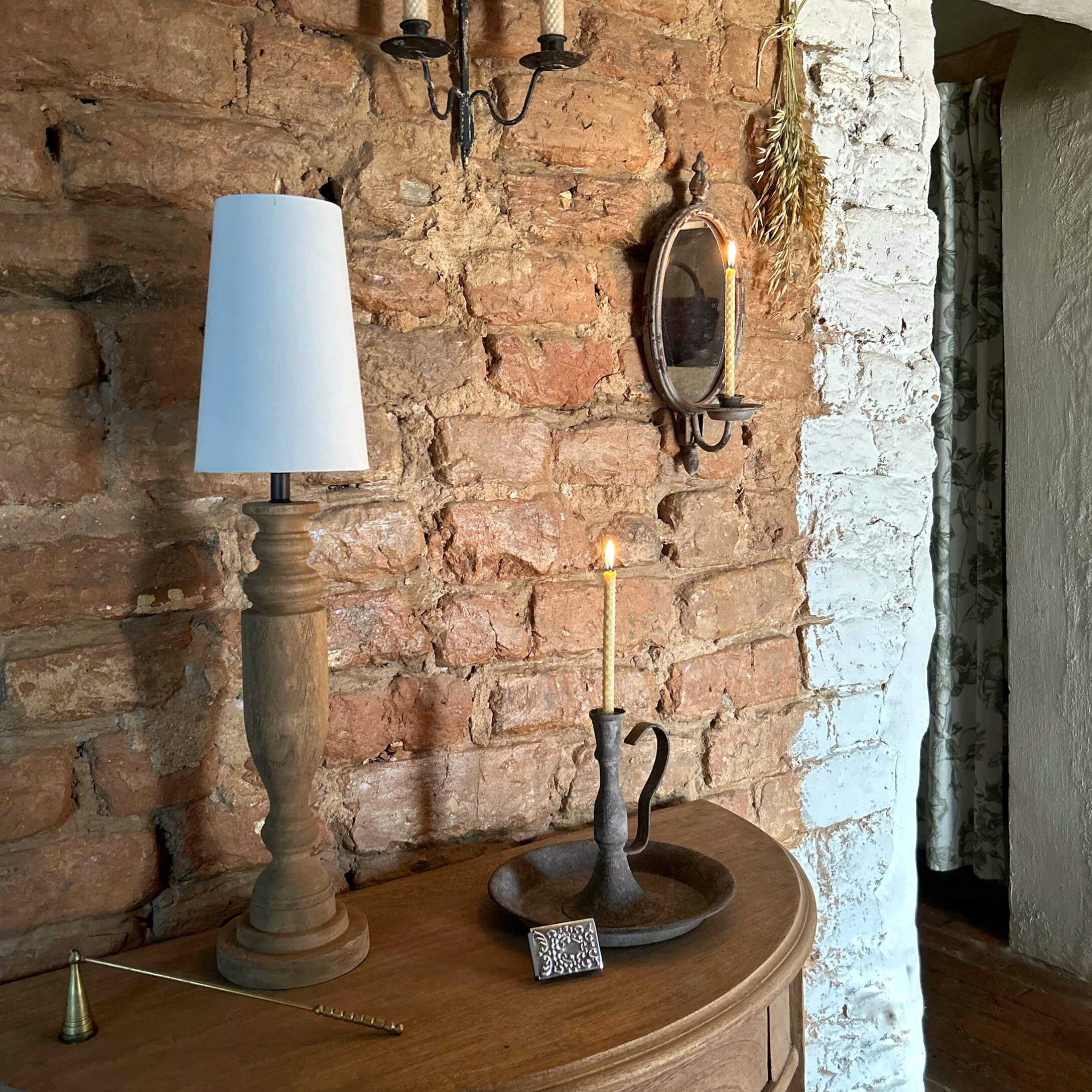 Holztisch mit Holzlampe mit weißem Schirm und Metallkerzenhalter mit brennender Kerze vor Ziegelwand