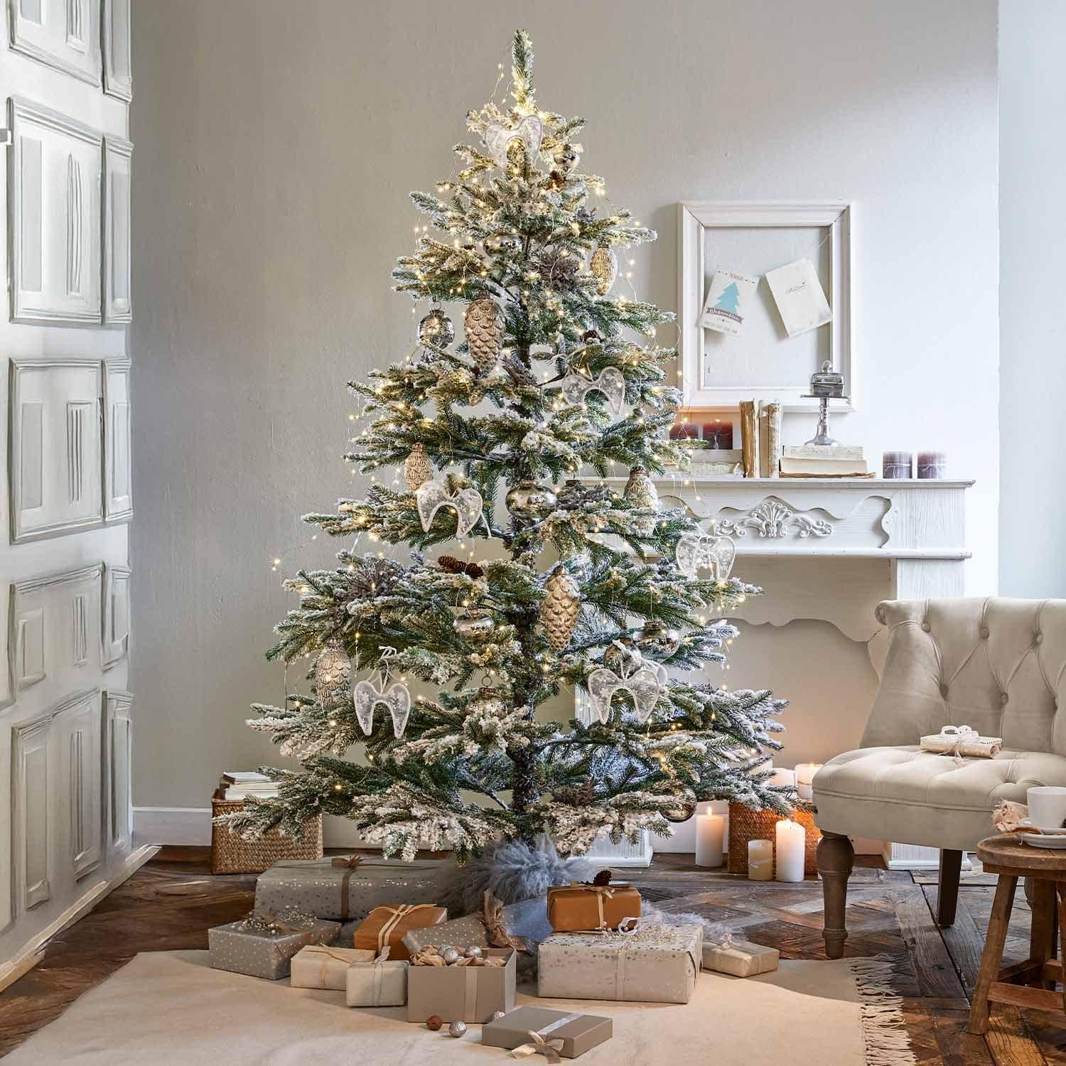 Schneebedeckter Weihnachtsbaum mit weißen Engelsanhängern und goldenen Zapfen in gemütlichem Wohnzimmer mit Kamin