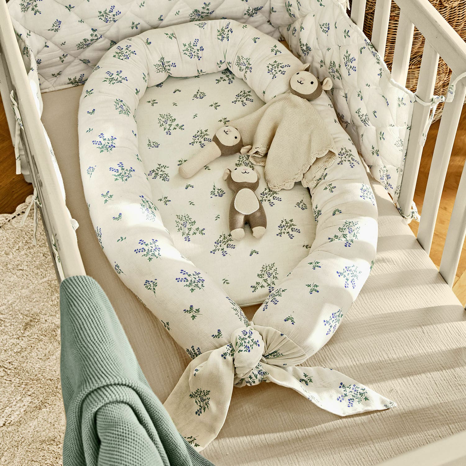 Baby-Nestchen Nymariel Weißes Baby-Nest aus Baumwolle mit blau-grünem Blumenmuster und Kuscheltieren im hellen Babybett