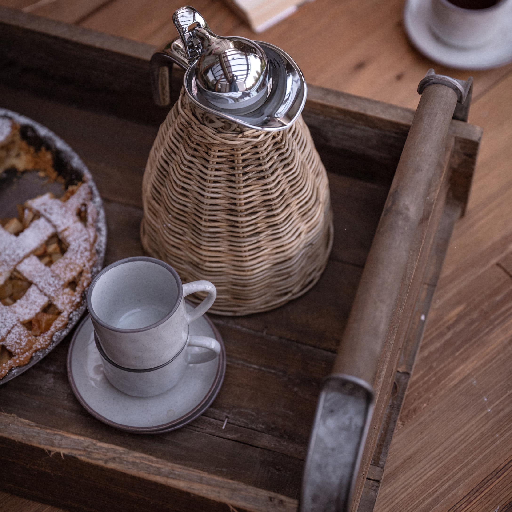 Holztablett mit geflochtenem Krug und zwei weißen Tassen neben Kuchen auf Holztisch