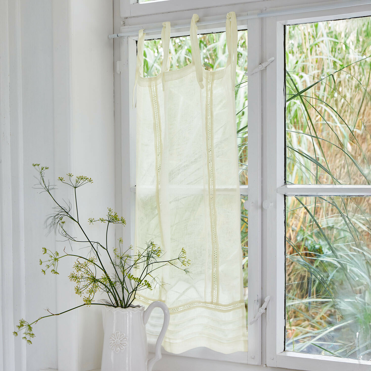 Cremefarbener Vorhang aus leichtem Stoff mit dekorativen Bändern an weißem Fenster, Wohnaccessoire