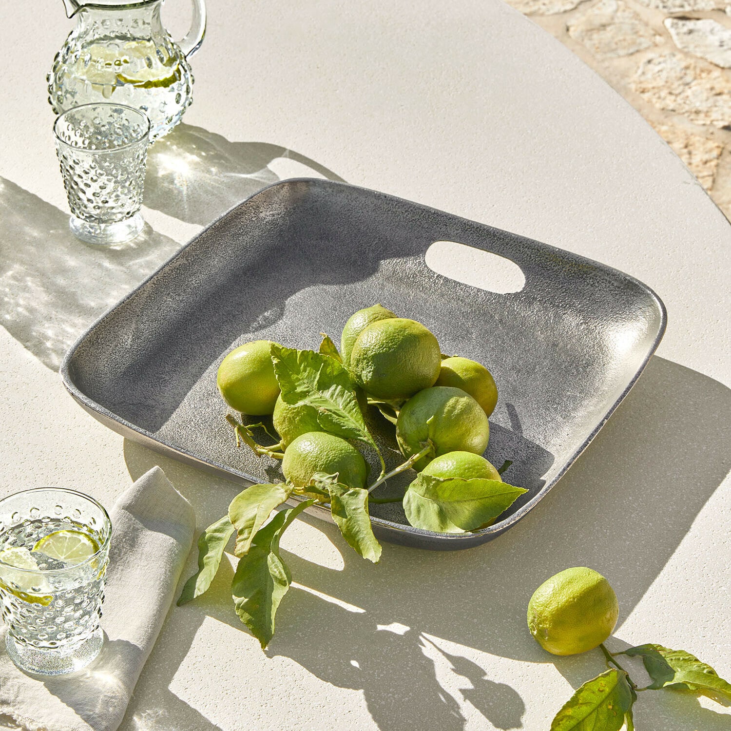 Quadratische graue Metallplatte mit grünen Limetten auf hellem Tisch, sommerliche Tischdekoration