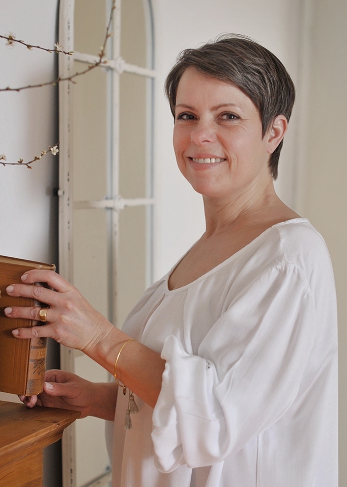 Frau in weißer Baumwollbluse hält altes Buch an einem Holzregal in hellem Raum