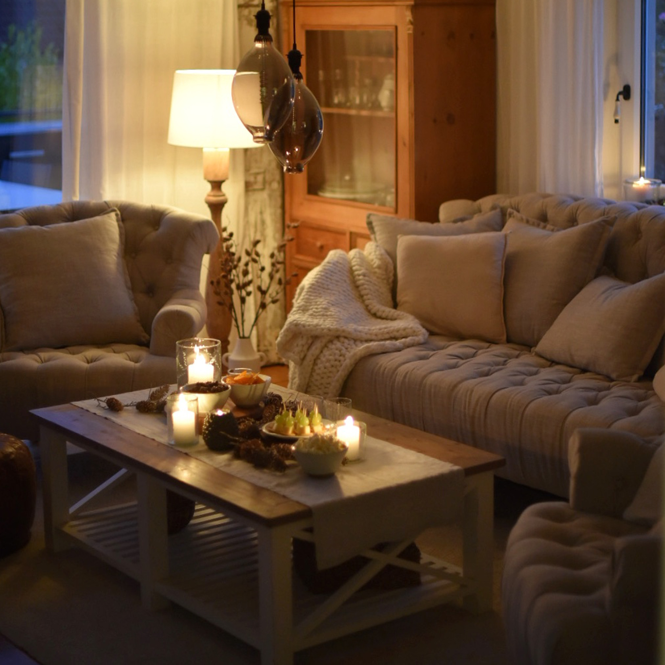 Gemütliches Wohnzimmer mit beigem Sofa, Sesseln und Holztisch mit Kerzen und Snacks dekoriert