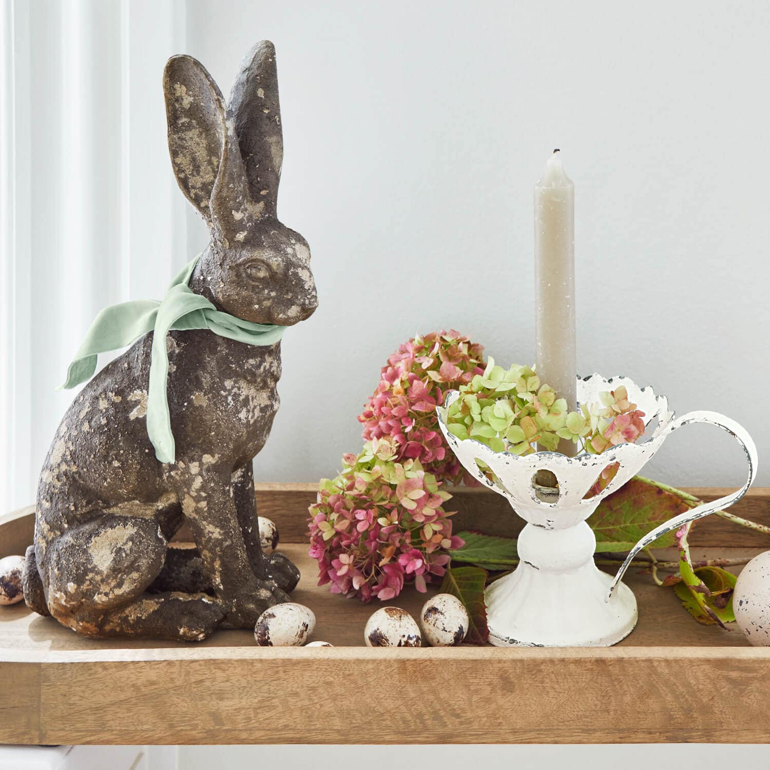Dekorativer Hase aus patiniertem Metall mit grünem Band, weißer Metallkerzenhalter und Blumen auf Holztablett