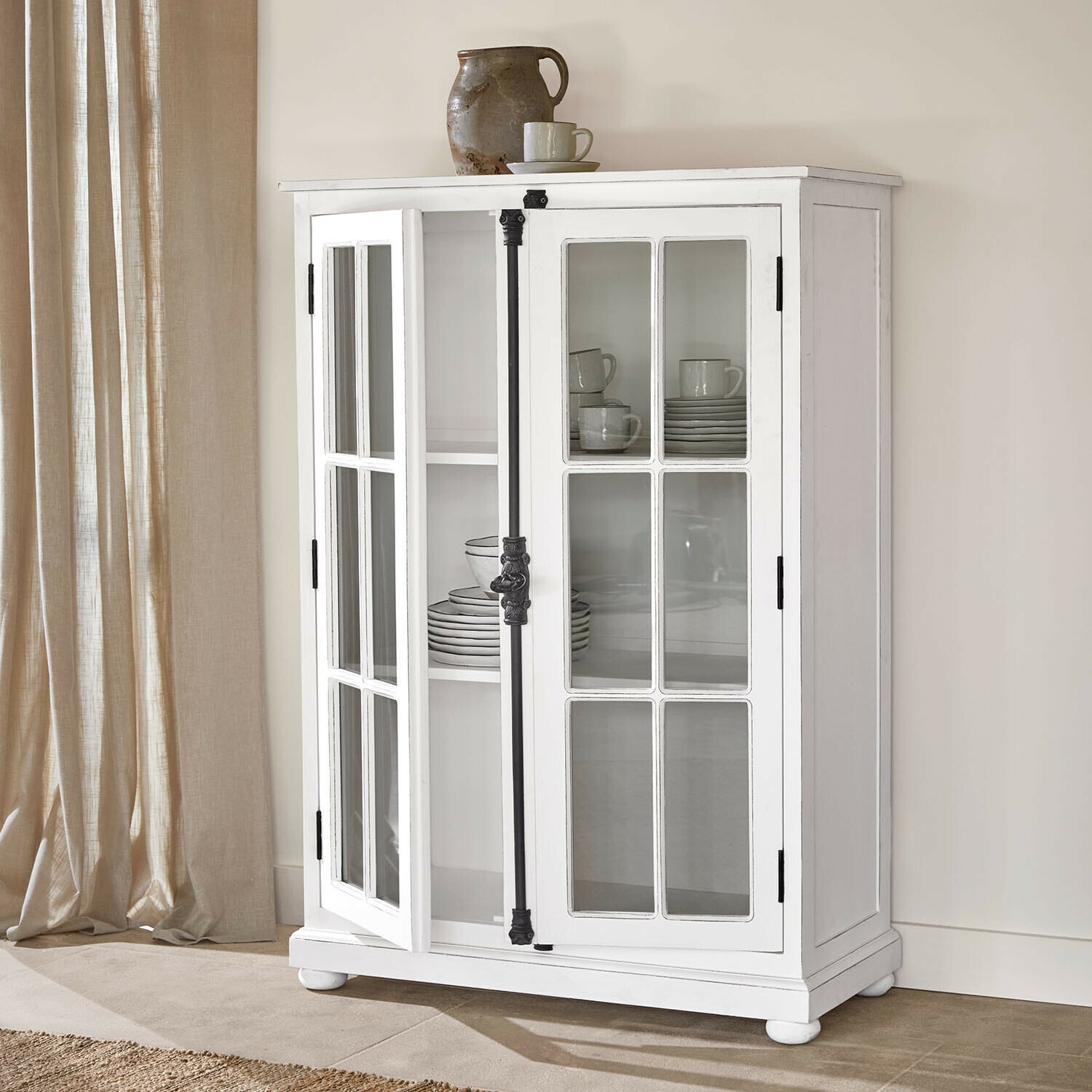 Schrank Salivonne Weiße Holzvitrine mit verglasten Türen und schwarzen Metallgriffen im Vintage-Stil in hellem Raum