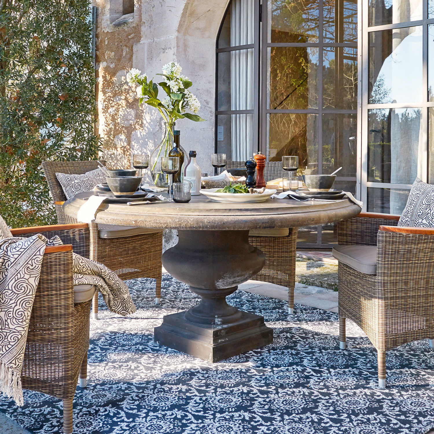 Tisch Monterey Runder Steintisch mit schwarzem Sockel und Rattanstühlen auf Terrasse mit gemustertem Teppich