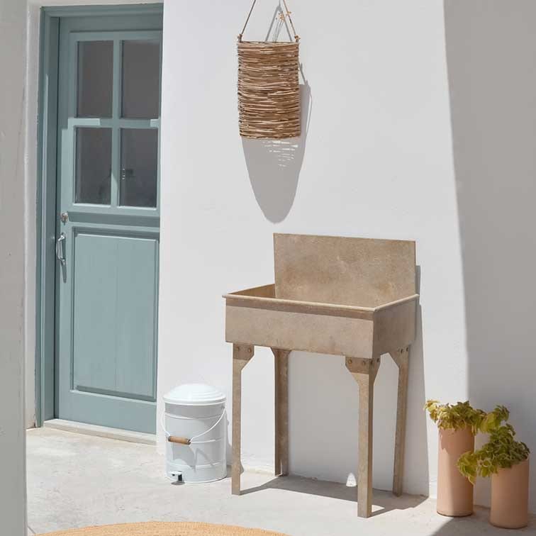 Beige Steintopf auf Beinen vor weißer Wand mit blauer Tür und hängendem Korb aus Weide