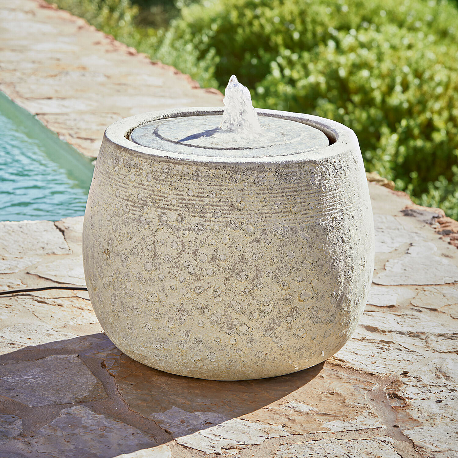 Runder Gartenbrunnen aus hellem Stein mit feinem Muster und Wasserstrahl auf Steinterrasse