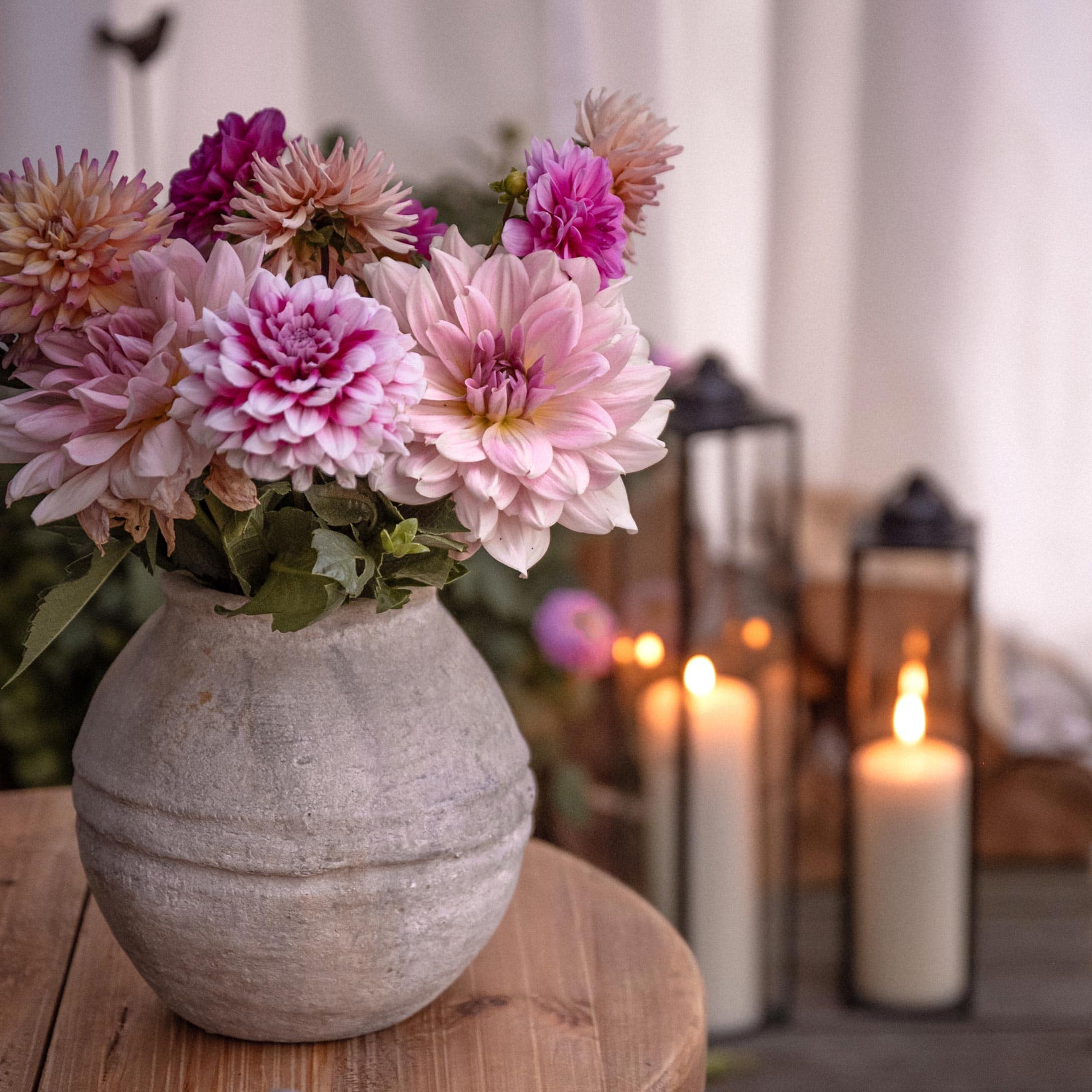 Grauer Dekovase mit rosa-violetten Blumen auf Holztisch, brennende Laternen im Hintergrund