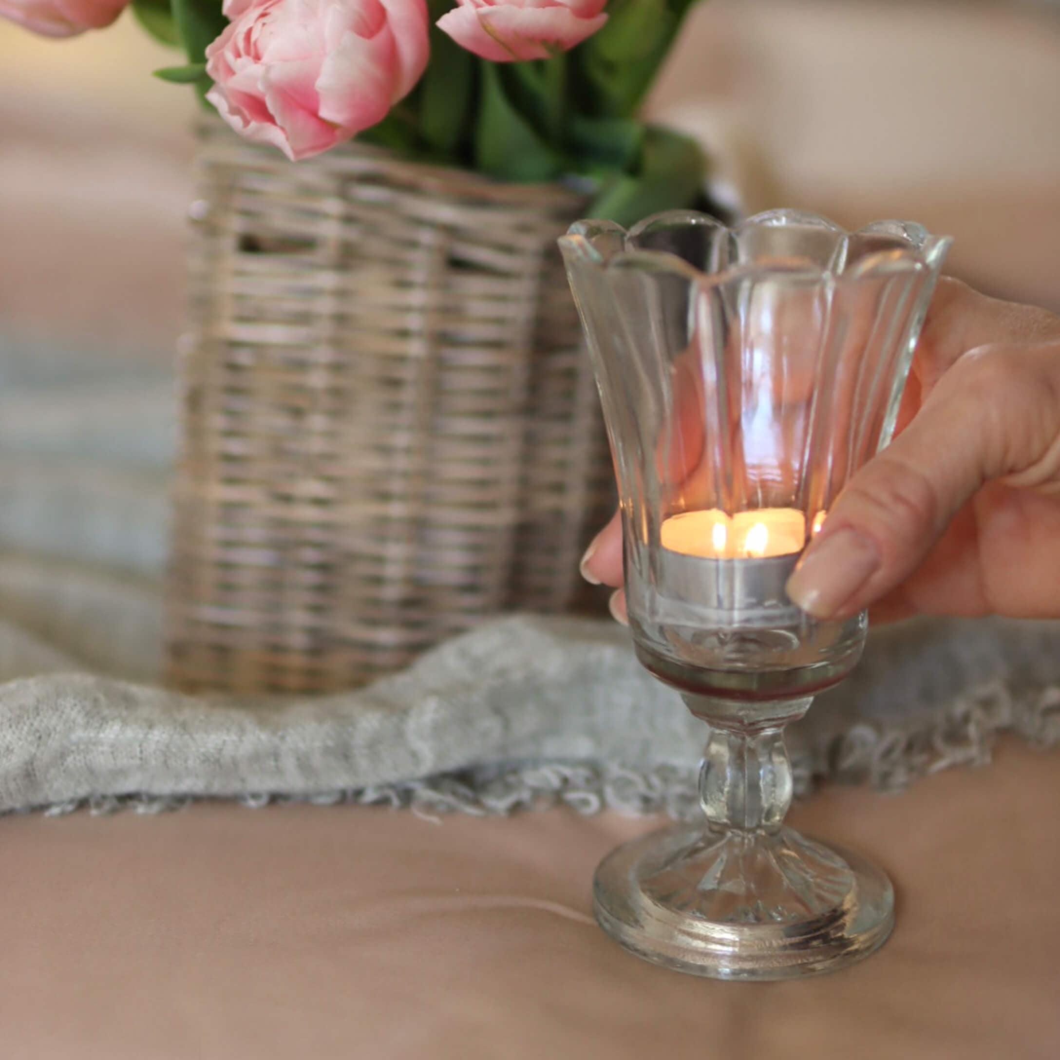 Glas-Teelichthalter auf Fuß mit brennender Kerze, im Hintergrund rosa Blumen in Korbvase