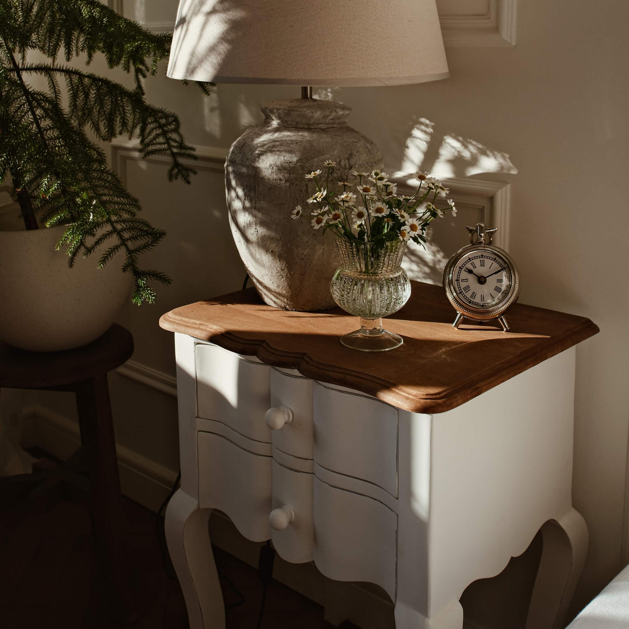 Weiße Holzkommode mit brauner Holzplatte, Lampe, Glasvase mit Blumen und silberner Uhr im Sonnenlicht
