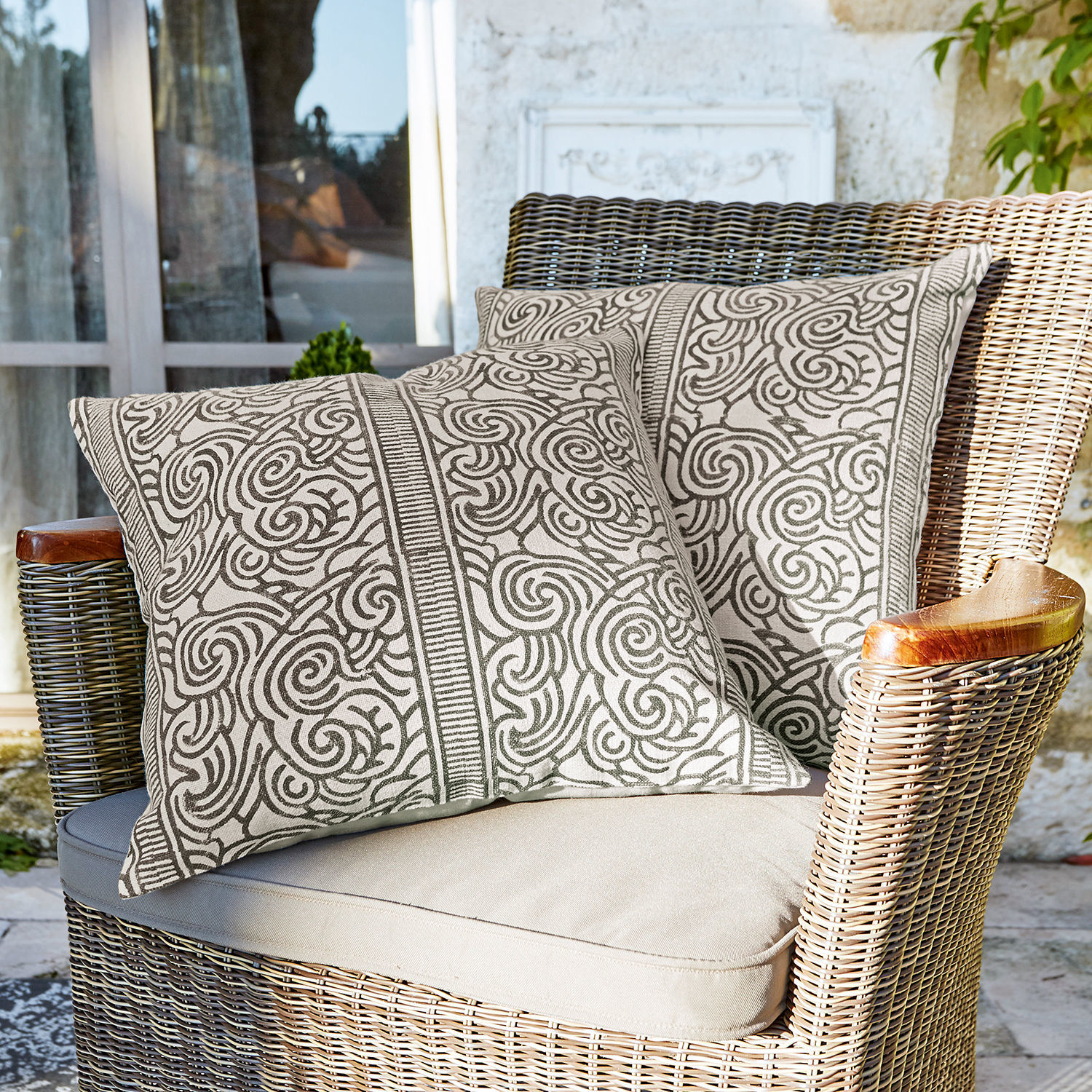 Kissenhülle 2er Set Ensis Zwei dekorative Baumwollkissen in Beige mit braunem Muster auf Rattansessel auf Terrasse